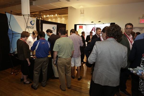 Attendees wearing lanyards crowd an exhibit hall near an Amcor booth and another exhibit with a bottle graphic on its banner.