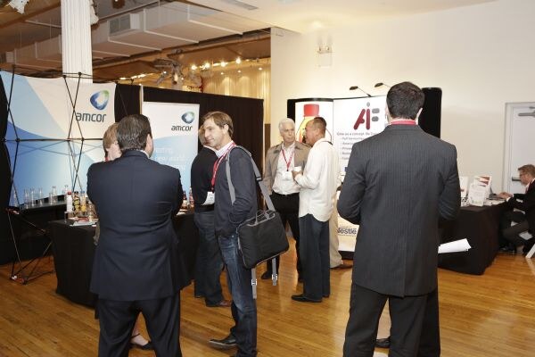 Attendees talk in an exhibit hall between booths. An Amcor banner and display stand on the left, and another booth with a partly visible banner stands at the back. Tables hold bottles and papers.