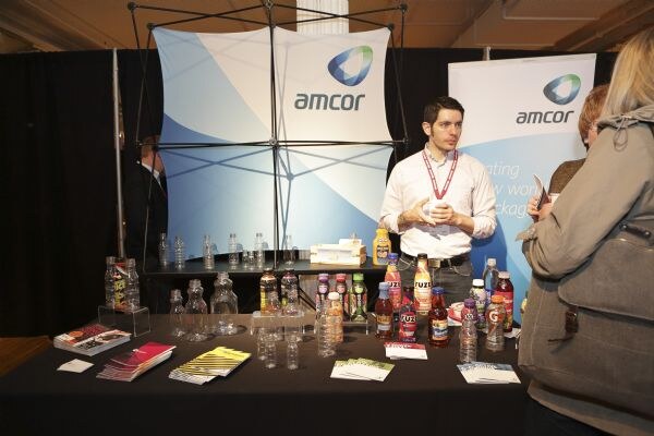 A man in a white shirt and lanyard stands behind a table of clear plastic bottles, filled beverage bottles and brochures at the Amcor booth, with Amcor banners behind him and a visitor in the foreground.