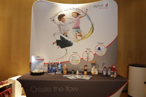 Aptar exhibit with a curved banner showing two children and a splash of liquid, above a table of bottles and brochures with the slogan "Create the flow" on the tablecloth.