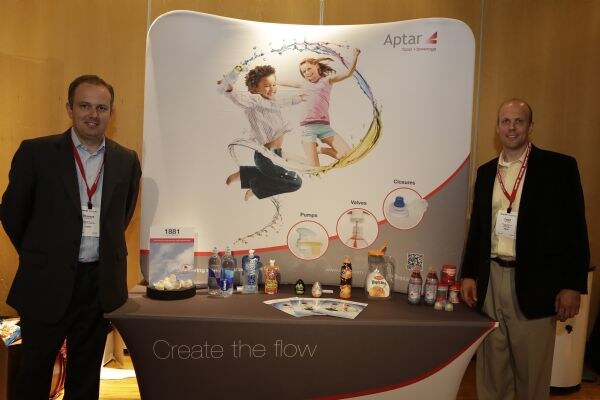 Two men in dark blazers and red lanyards stand on either side of the Aptar booth, which has a banner of two children, bottles and brochures on a table, and "Create the flow" on the cloth.