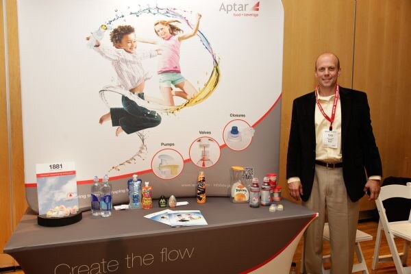 A man in a dark blazer and red lanyard stands beside the Aptar exhibit, which has a banner of two children, a table of bottles and brochures, and "Create the flow" on the cloth.