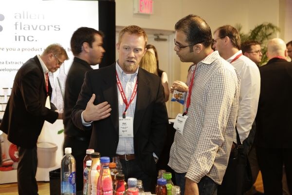 A bearded man in a dark jacket gestures while talking with a man in glasses and a checked shirt, beside a table of bottled drinks, with an Allen Flavors banner and other attendees behind them.
