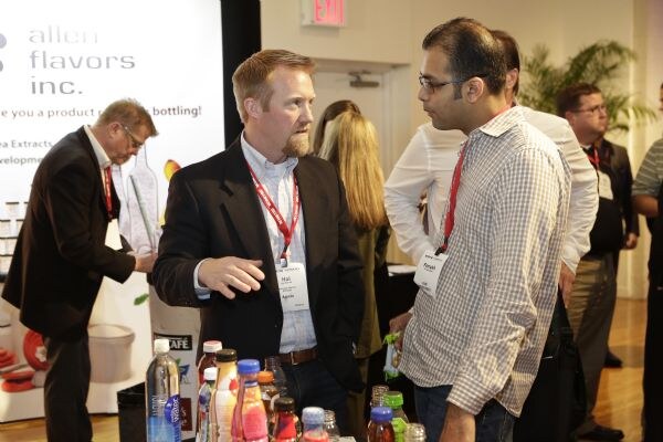 A bearded man in a dark jacket gestures while talking with a man in glasses and a checked shirt, with bottled drinks in front and an Allen Flavors Inc. banner behind them.