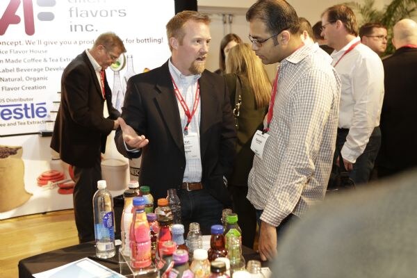 A bearded man in a dark jacket gestures toward a man in glasses and a checked shirt over a table of bottled drinks, with Allen Flavors Inc. and Nestle banner text and other attendees behind them.