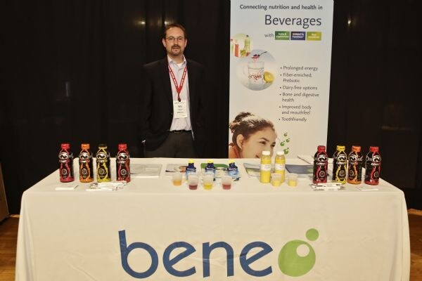A man with glasses and a red lanyard stands behind a white Beneo tablecloth set with bottles, sample cups and brochures, in front of a banner headed "Beverages."