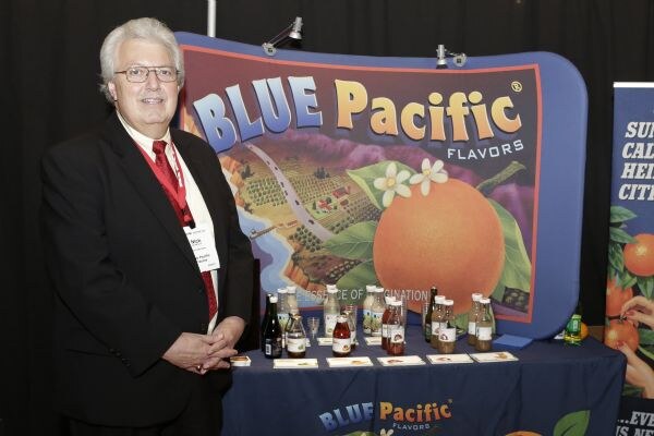 A gray-haired man with glasses, a red tie and a dark suit stands beside a Blue Pacific Flavors display. The table in front of the orange-themed banner holds bottles and cards.