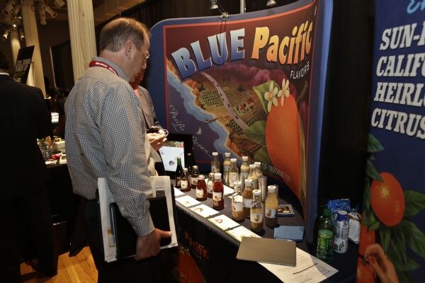 A man in a checked shirt holding papers looks at bottles and cards on the Blue Pacific Flavors table, which sits below an orange-themed banner.