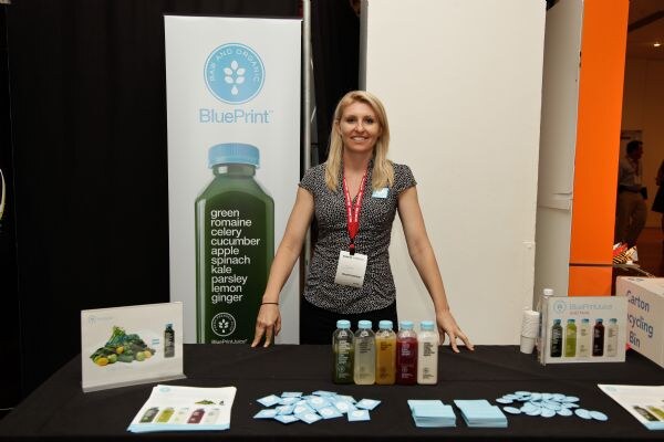 A blonde woman with a lanyard stands behind a black-draped BluePrint table holding five juice bottles, brochures and small blue cards, in front of a BluePrint banner showing a green juice bottle.