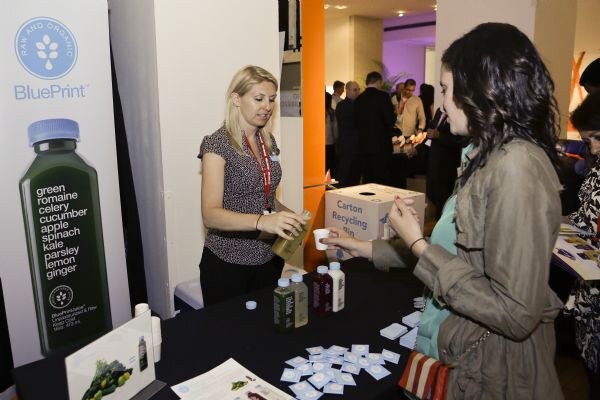 A blonde woman wearing a lanyard stands behind a BluePrint table and hands a small cup to a dark-haired woman in a gray jacket. A BluePrint banner and juice bottles are visible.