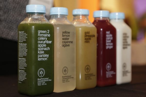 Five BluePrintJuice bottles with blue caps in a row, filled with green, pale yellow, gold, deep red and white juices. Labels list ingredients such as romaine, celery, lemon and pineapple.