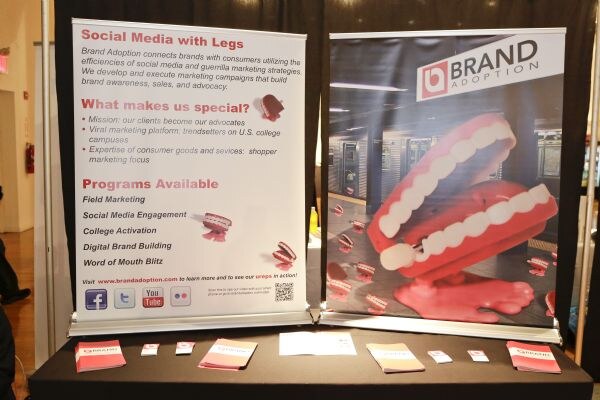 Two Brand Adoption banners on a table. One lists social media marketing programs; the other shows the Brand Adoption logo over red wind-up chattering teeth. Brochures lie on the table.