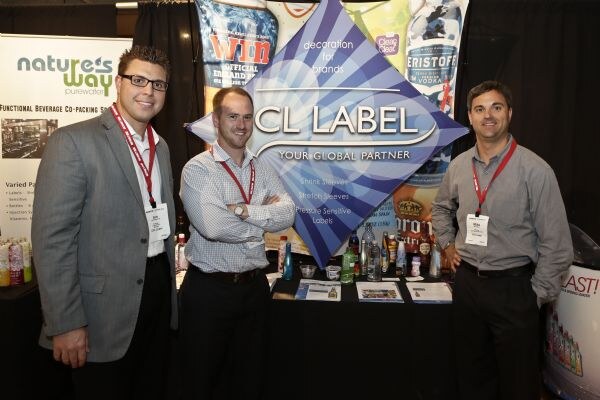 Three men in lanyards stand in front of a CCL Label diamond-shaped sign and a Nature's Way Purewater banner, with sample bottles and brochures on the table between them.