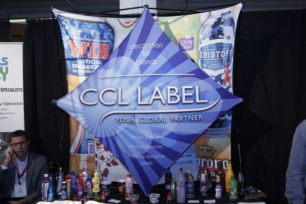 A CCL Label diamond sign reading 'Your Global Partner' hangs before a backdrop showing Eristoff and Clean & Clear packaging, with sample bottles on the table. A man on a phone sits at left.