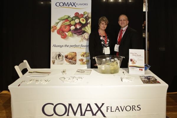 A woman and a man in lanyards stand behind a white-draped Comax Flavors table with a glass bowl, brochures and a banner showing fruits and vegetables and the line 'Always the perfect flavor'.