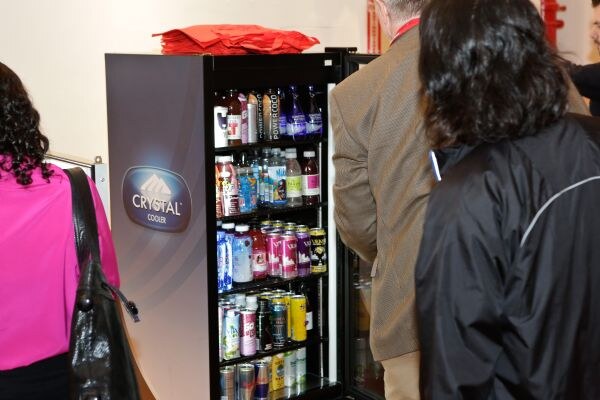 A black Crystal Cooler display refrigerator stocked with bottled and canned drinks, with two attendees standing beside it and a stack of red bags on top.