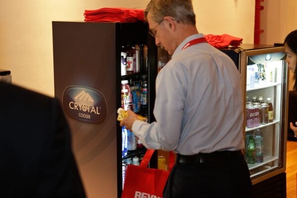 A man in a light blue shirt and red lanyard reaches into a Crystal Cooler stocked with drinks, holding a red bag. A second glass-door cooler stands beside it.