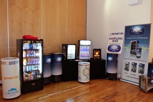 Crystal Cooler display of glass-door refrigerators and round coolers, one marked BevNET Live Summer 2012, beside a sampling bar sign and a Crystal Cooler banner on a wood floor.