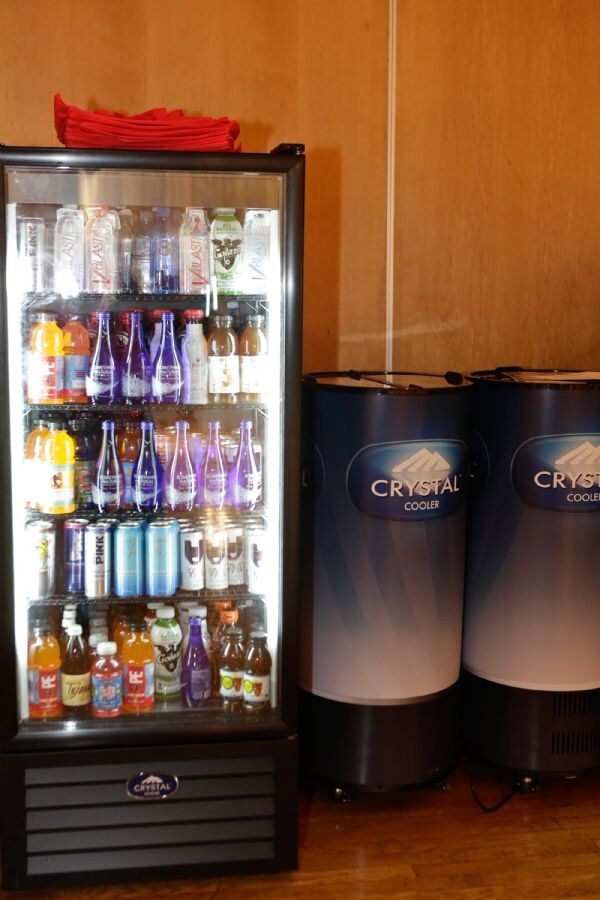 A glass-door Crystal Cooler refrigerator stocked with bottles and cans, beside two round Crystal Cooler coolers against a wood-paneled wall.