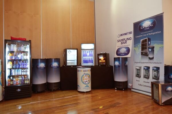 Crystal Cooler refrigerators and cylindrical coolers stand on a wood floor near a BevNET Live sampling bar sign and a Crystal Cooler banner. A lit glass-door cooler holds bottled drinks at left.