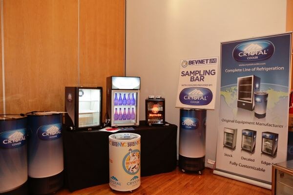 Crystal Cooler exhibit with drum coolers, small countertop coolers on a black-draped table, a BevNET Live Sampling Bar sign and a Crystal Cooler banner showing its line of refrigeration.