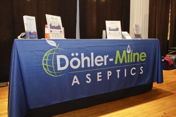 Table draped in a blue Döhler-Milne Aseptics cloth with brochures and signs on top, set on a wood floor in front of dark curtains.