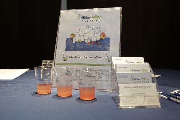 Table with three small cups of orange-pink drink in front of a Döhler-Milne Aseptics Blueberry Coconut Water sign, with business cards in holders and pens on a blue tablecloth.