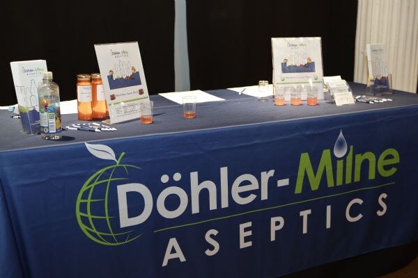 Table with a blue Döhler-Milne Aseptics cloth holding sample cups, bottles, jars, pens, brochures and framed signs.