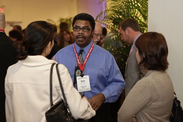 Donn, in a blue shirt, patterned tie and red lanyard with a name badge, talks with attendees. Two women stand in the foreground with their backs partly turned, and other people are behind him.