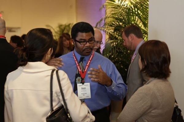 Donn, in a blue shirt, tie and red lanyard with a name badge, gestures with one hand while speaking. Two women face him in the foreground, and people and plants are behind.