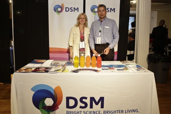A woman and a man stand behind a DSM table with five bottles of colored drinks and brochures, in front of DSM banners.