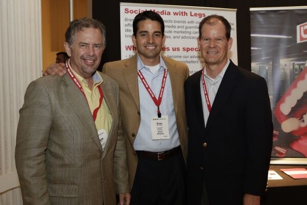 Three smiling men in jackets and lanyards pose together. Evan of Brand Adoption, per his badge, stands in the center in a tan blazer. A Brand Adoption banner is behind them.