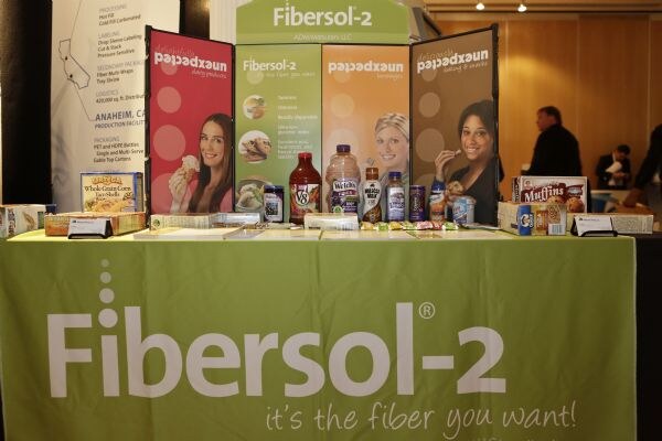 Green Fibersol-2 tablecloth with product displays, including bottled drinks, a V8 bottle, boxes and muffins, in front of panels showing smiling women.