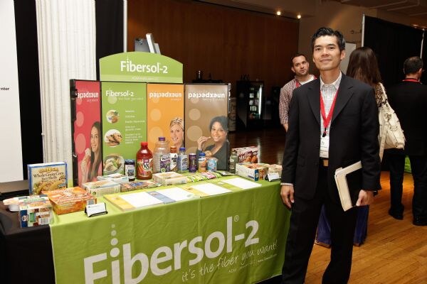 A man in a dark suit with a lanyard stands beside a Fibersol-2 table of food and drink products, with other attendees behind him.