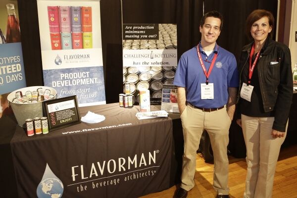 A man in a blue polo and a woman in a black jacket, both wearing lanyards, stand beside a table with a Flavorman banner. A Challenge Bottling sign is behind them.