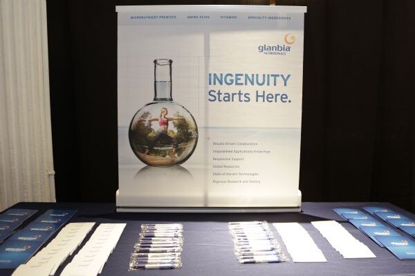 Close view of a Glanbia Nutritionals banner reading "Ingenuity Starts Here," above a table with brochures and pens.