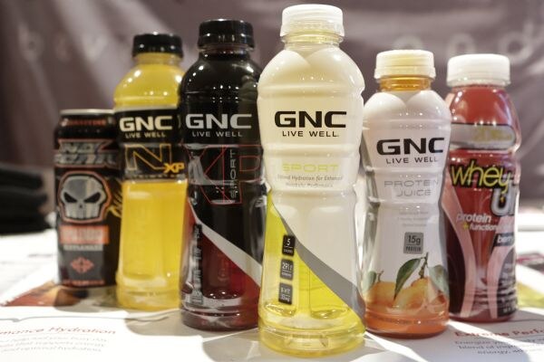 Six beverage containers in a row, including GNC Live Well sport drinks and protein juice, a GNC bottle with yellow drink and a dark can with a skull design.
