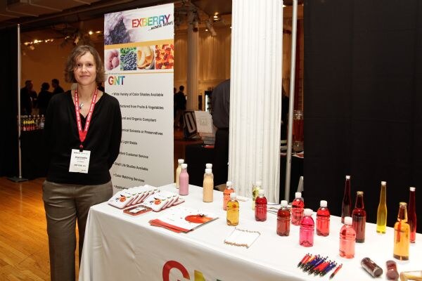 A woman with a lanyard stands beside a GNT Exberry banner and a table of colorful liquid samples in bottles, with brochures and pens.