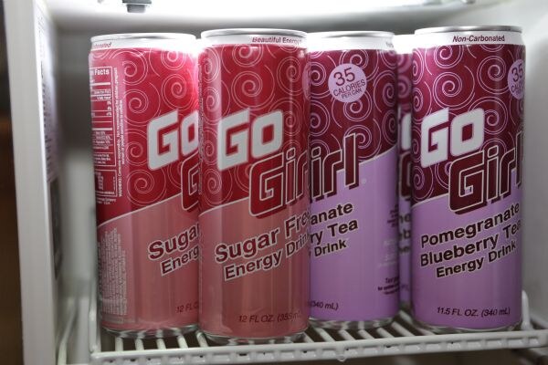 Four tall cans of Go Girl energy drink on a cooler shelf, two red Sugar Free cans and two purple Pomegranate Blueberry Tea cans marked 35 calories per can.