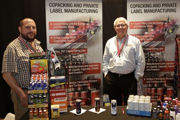 Two men with red lanyards stand at a table in front of copacking and private label manufacturing banners, beside a display of I Am Energized shots and cans of Mercy.