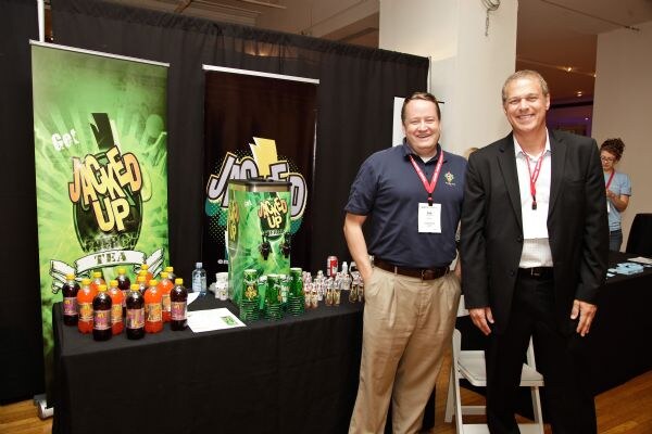 Two smiling men wearing lanyards stand beside a table with Jacked Up tea bottles and a dispenser. Green and black Jacked Up banners are behind them.
