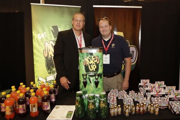 Two men with lanyards stand behind a table with a Jacked Up energy dispenser, green cups, bottled drinks and shot displays, in front of Jacked Up banners.