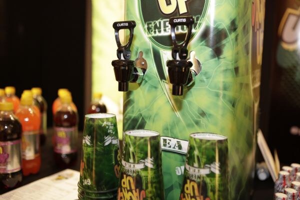 A green Jacked Up energy dispenser with two Curtis spigots and stacked green Jacked Up cups in front, with bottled drinks on the table at left.
