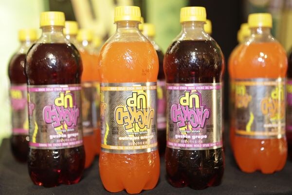 Plastic bottles of Jacked Up energy drink with yellow caps in Gushin Grape and Outrajus Orange flavors, with upside-down labels, on a dark table.