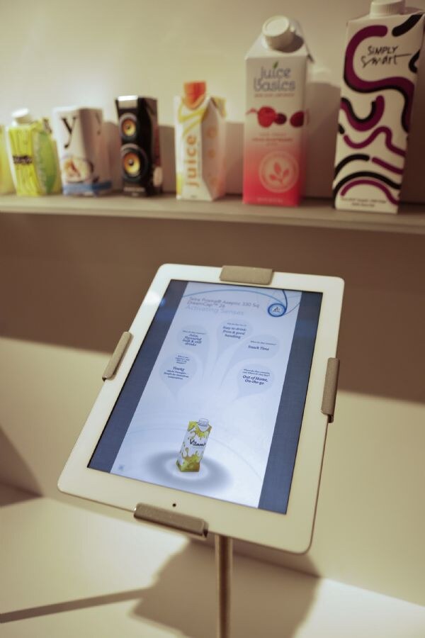 A tablet on a stand in the foreground shows a Tetra Pak screen. On a shelf behind it are cartons, including Juice Basics and Simply Smart, beside a speaker-shaped carton.
