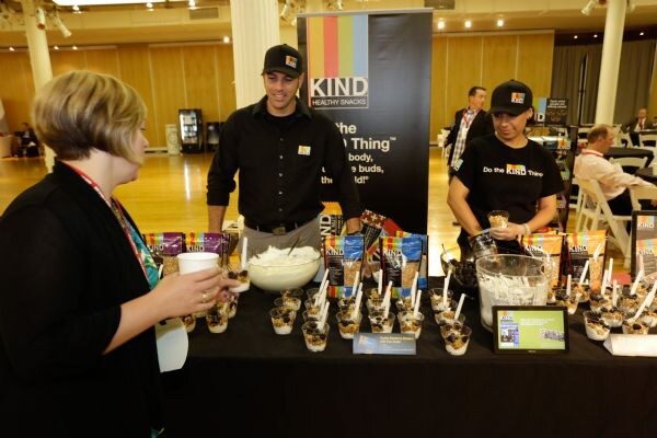 Two people in black KIND shirts and caps stand behind a table of snack cups and KIND packages as a woman in black holds a cup in the foreground.