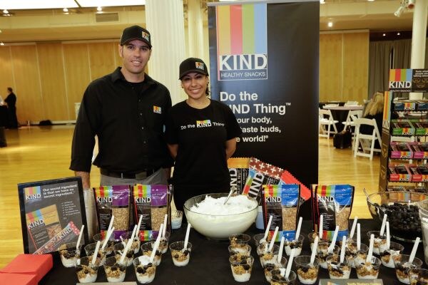A man and a woman in black KIND shirts and caps stand behind a table of snack cups, KIND packages and a bowl, with a banner and a KIND display rack behind.