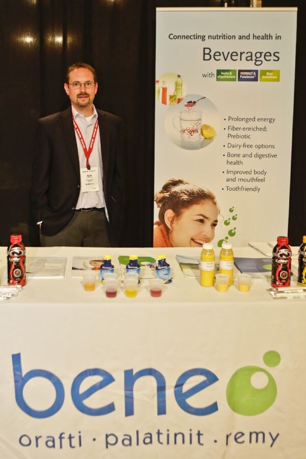 Kyle, a man with glasses and a red lanyard, stands behind the Beneo table. The table has bottles, sample cups and brochures, and a banner about beverage nutrition stands behind him.