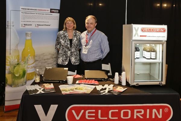 A woman and a man stand behind a black table with Velcorin branding, brochures and a small Velcorin refrigerator, beside a Lanxess banner.
