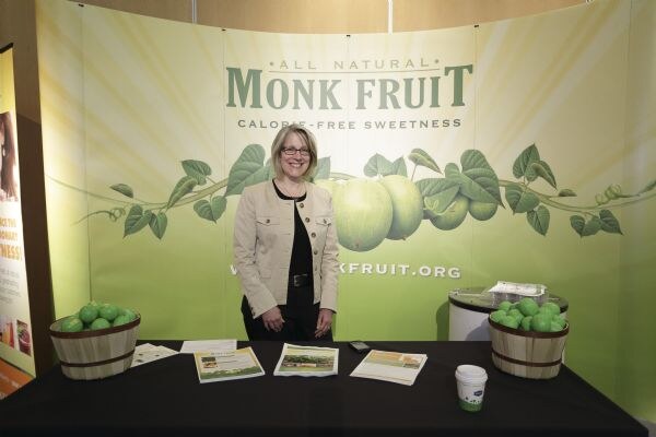 A smiling woman with glasses and a tan jacket stands behind a table at the Monk Fruit booth. Behind her is a green banner reading All Natural Monk Fruit, with baskets of green fruit and brochures on the table.